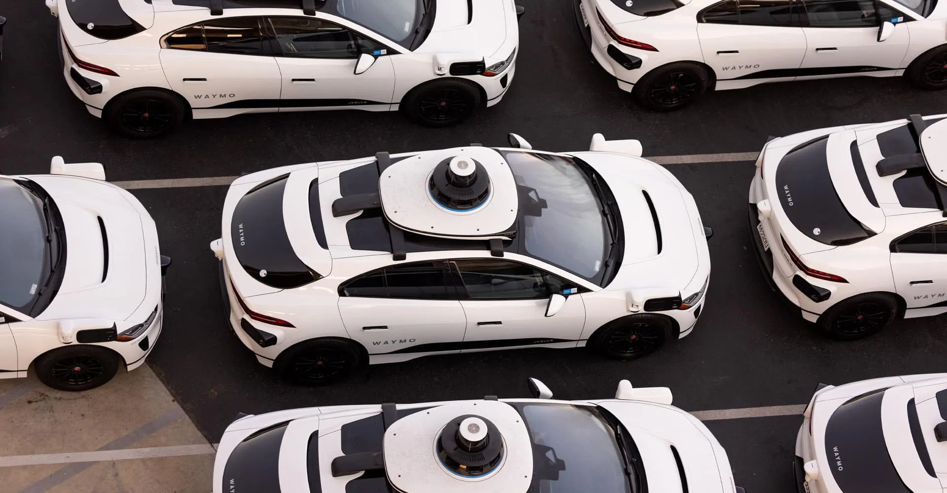 Overhead view of a parked fleet of white Waymo self-driving Jaguar I-Pace cars, each with a roof-mounted lidar sensor.