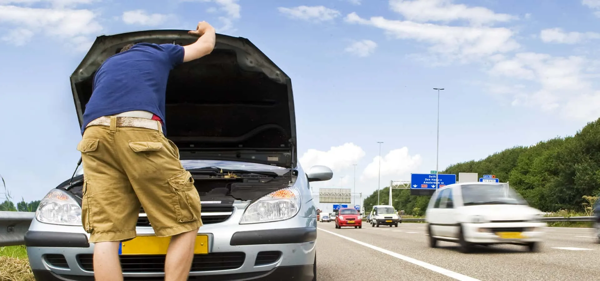 A driver leans under the open bonnet of a broken-down car stopped on the shoulder of a motorway as traffic passes in the next lane.