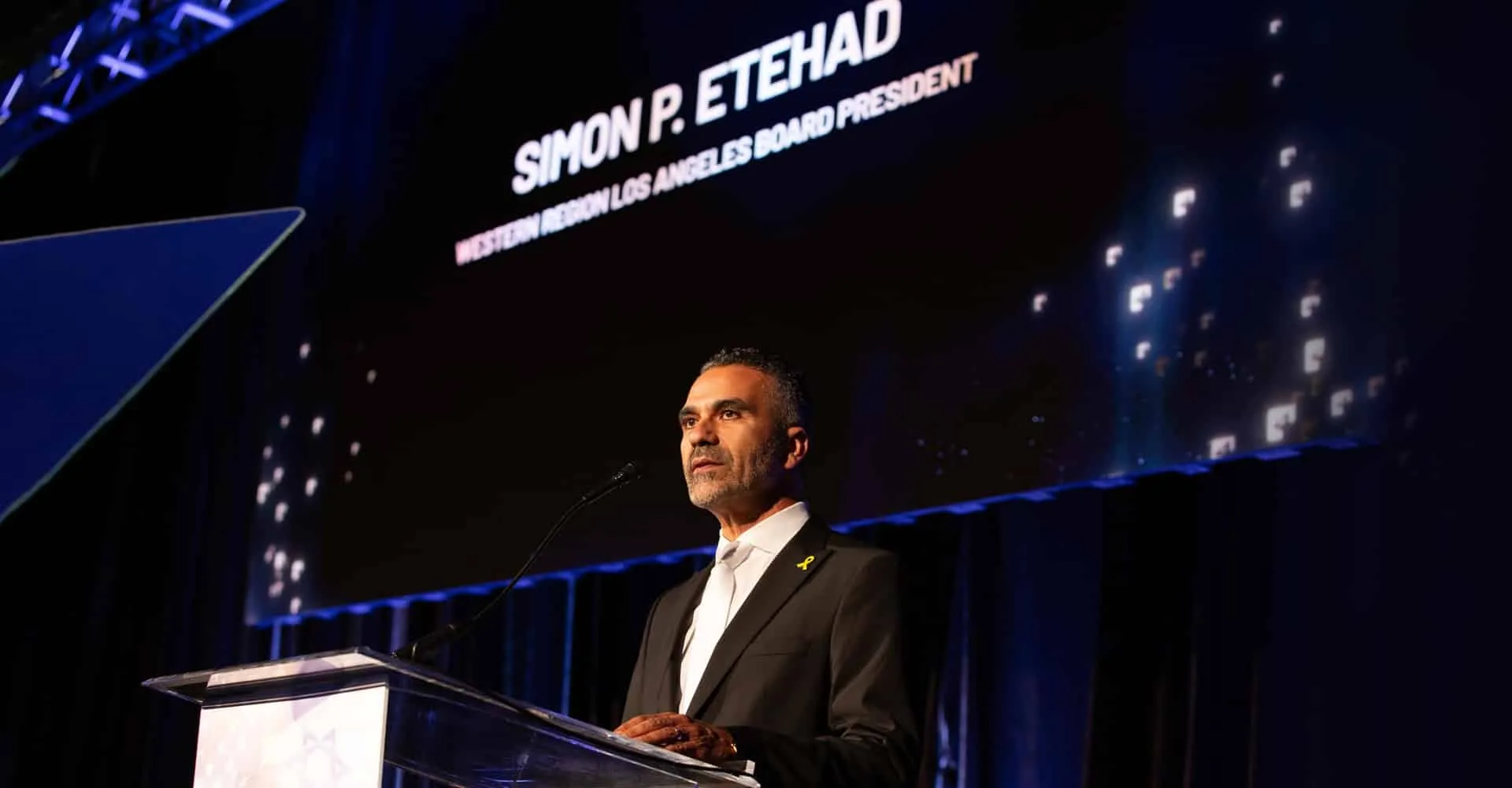 Simon Etehad speaking at a lectern, with a screen behind him reading "Simon P. Etehad, Western Region Los Angeles Board President".