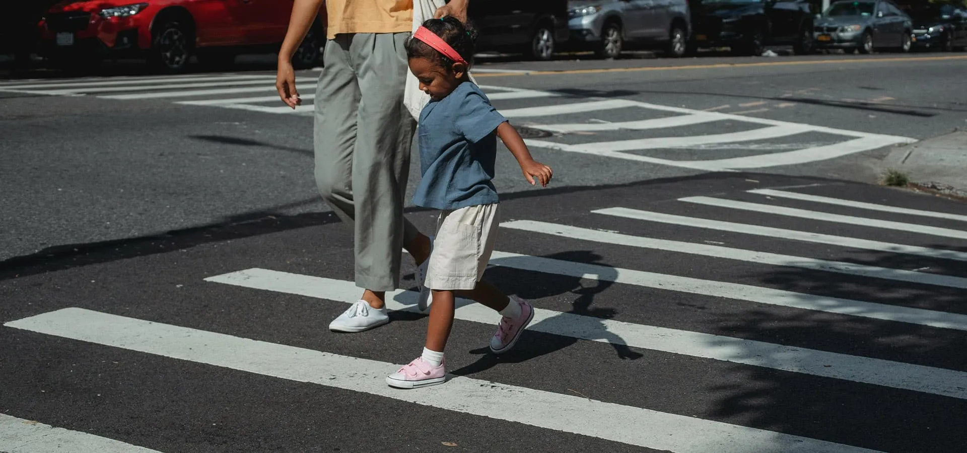 An adult holds the hand of a young child as the two of them cross a street inside a marked pedestrian crosswalk.